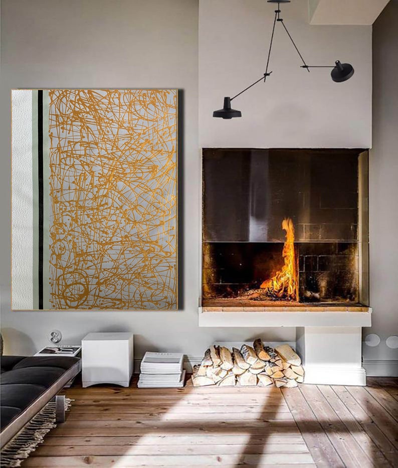 K&ouml;nnte beinhalten: Ein modernes Wohnzimmer mit einem Kamin und einem gro&szlig;en abstrakten Gem&auml;lde mit goldenen Linien auf wei&szlig;em Hintergrund. Das Gem&auml;lde h&auml;ngt an einer wei&szlig;en Wand. Der Kamin ist aus Ziegeln gebaut und hat einen schwarzen Metallrahmen. Vor dem Kamin liegt ein Stapel Brennholz.