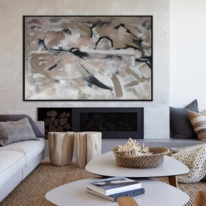 May include: Abstract painting in shades of brown, pink, and white with black accents. The painting is framed in a black frame and is hanging on a wall above a fireplace. The fireplace is made of stone and has a black mantel. There are two wooden stools in front of the fireplace.