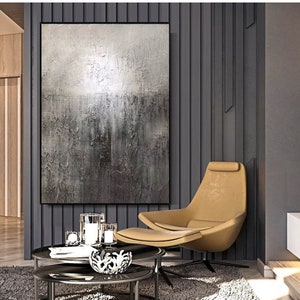 May include: A large abstract painting in shades of gray and white hangs on a wall in a modern living room. The painting is textured and has a subtle shimmer. A tan armchair with a footrest sits in front of the painting. Two glass coffee tables are in front of the chair.