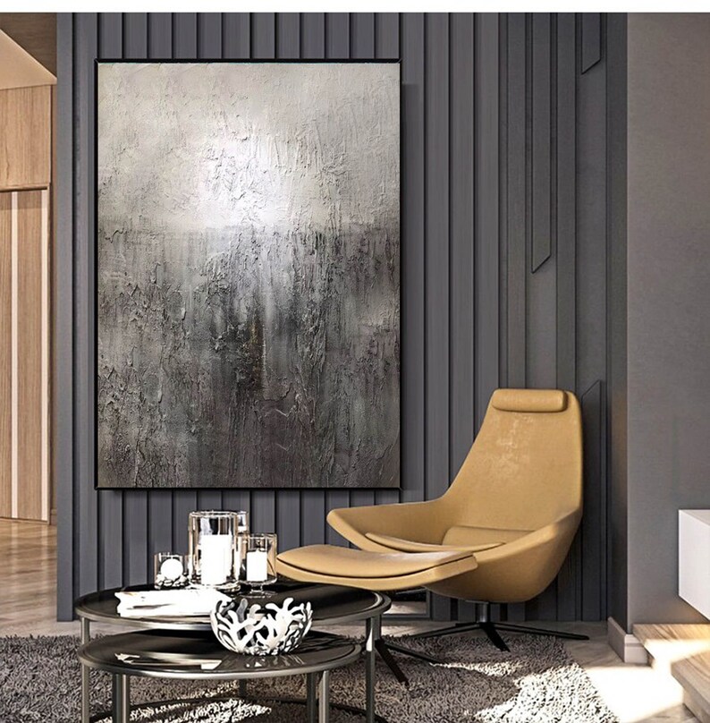 May include: A large abstract painting in shades of gray and white hangs on a wall in a modern living room. The painting is textured and has a subtle shimmer. A tan armchair with a footrest sits in front of the painting. Two glass coffee tables are in front of the chair.