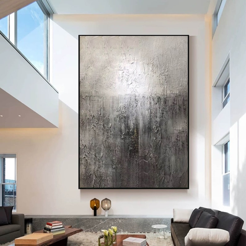 May include: A large abstract painting in shades of gray and white with a textured surface. The painting is framed in black and hangs on a white wall in a modern living room.