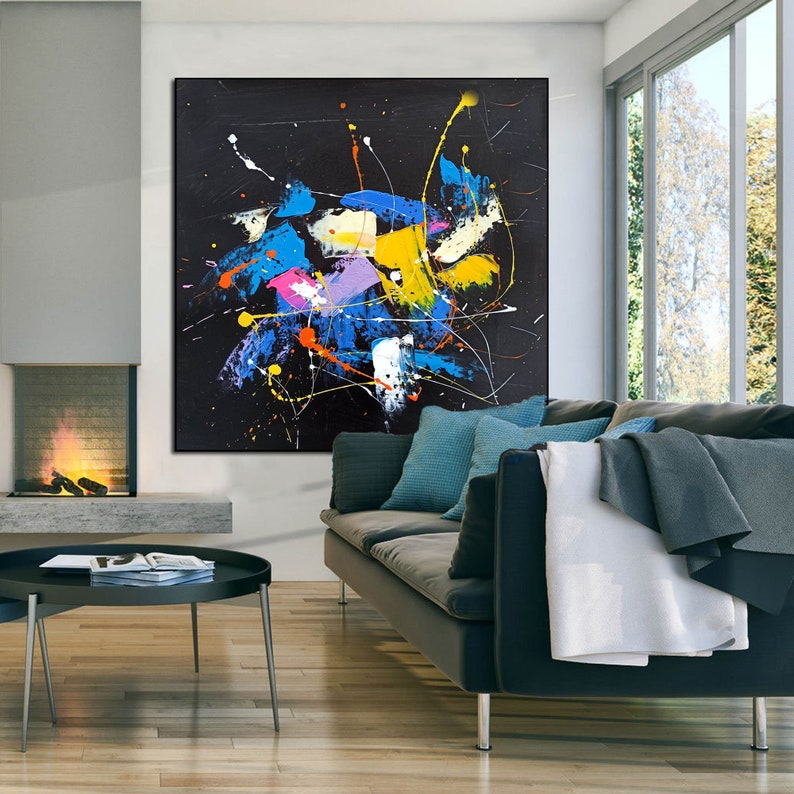 May include: Abstract painting with splashes of blue, yellow, pink, and white paint on a black background. The painting is hanging on a wall in a living room setting.