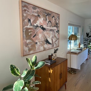 May include: A mid-century modern cabinet with two doors and a light wood finish. The cabinet is decorated with a potted plant and a framed abstract painting with pink, brown, and white colors.