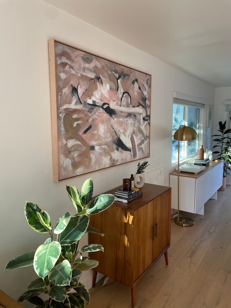 May include: A mid-century modern cabinet with two doors and a light wood finish. The cabinet is decorated with a potted plant and a framed abstract painting with pink, brown, and white colors.