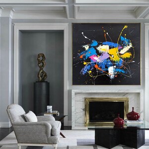 May include: A modern living room with a fireplace and a large abstract painting above it. The painting features a black background with splashes of blue, yellow, pink, and orange paint. A gray armchair sits in front of the fireplace, and a glass coffee table sits in front of the armchair.