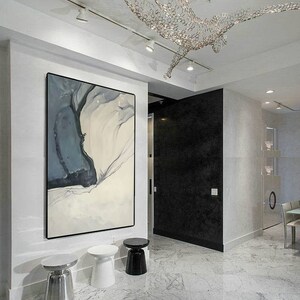 May include: A white, gray, and black abstract painting hangs on a white wall in a modern home. Three small round side tables are in the foreground. The tables are silver, white, and black.