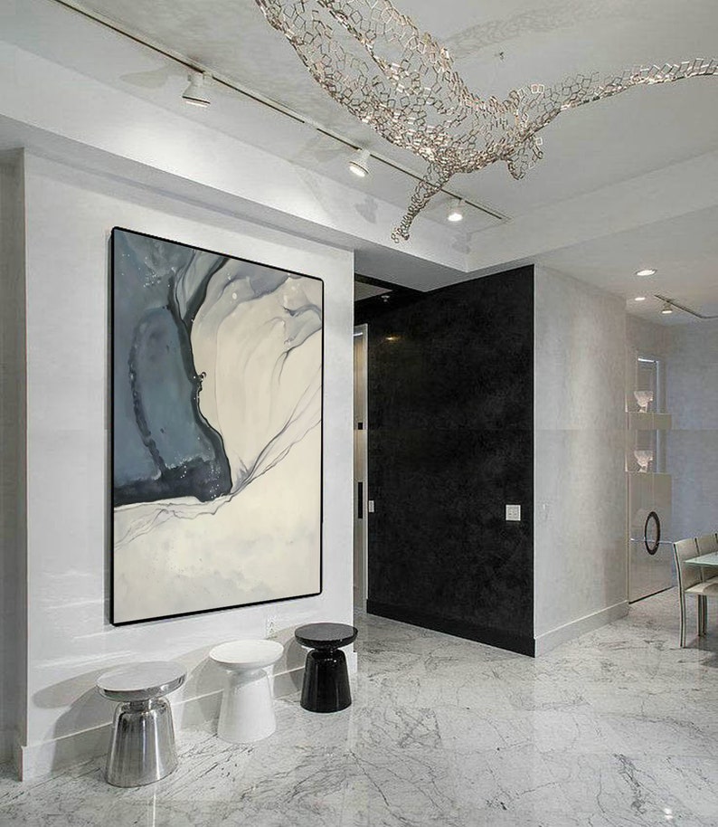 May include: A white, gray, and black abstract painting hangs on a white wall in a modern home. Three small round side tables are in the foreground. The tables are silver, white, and black.