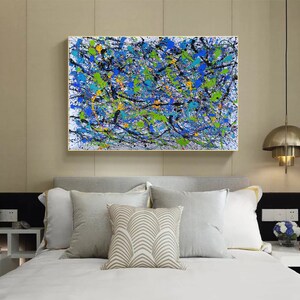 May include: Abstract painting with a white background and splashes of blue, green, black, and yellow paint. The painting is framed in a gold frame and is hanging on a wall above a bed with white bedding and pillows.