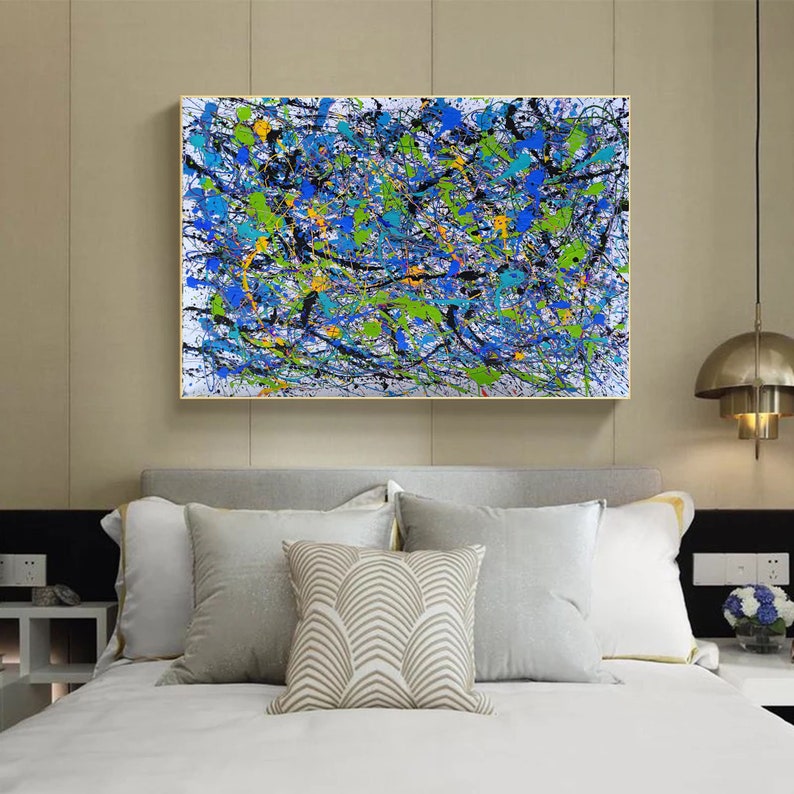 May include: Abstract painting with a white background and splashes of blue, green, black, and yellow paint. The painting is framed in a gold frame and is hanging on a wall above a bed with white bedding and pillows.
