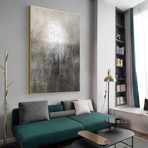 May include: A large abstract painting with a textured, metallic finish in shades of gray and silver. The painting is framed in a gold frame and hangs on a gray wall above a green sofa.