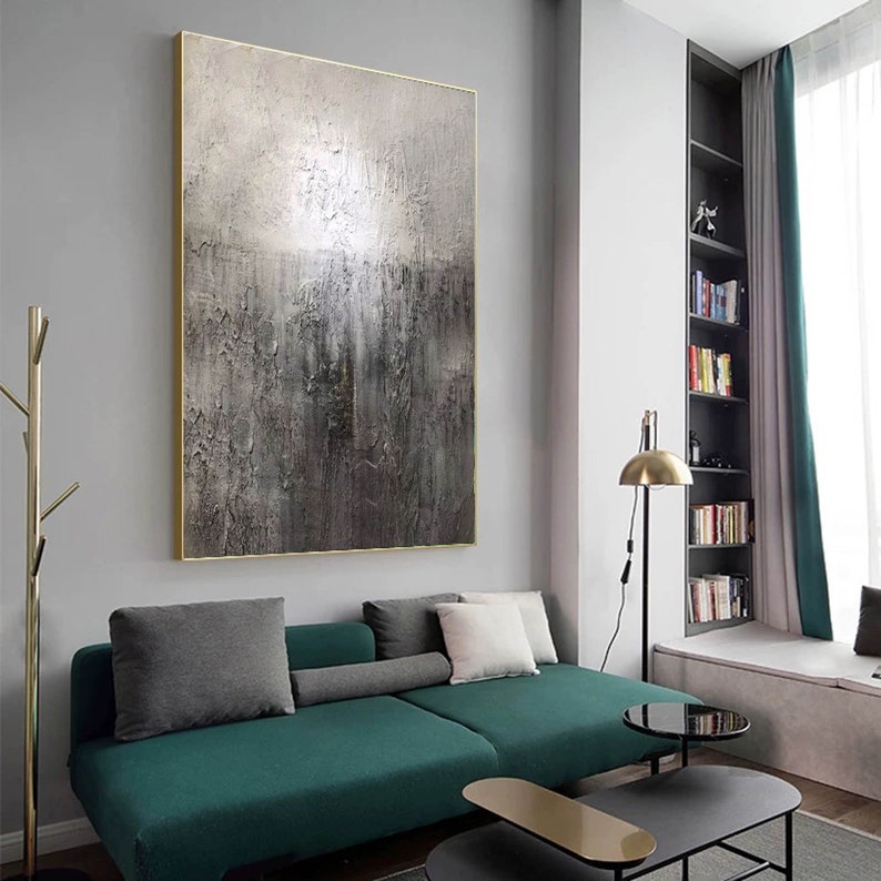 May include: A large abstract painting with a textured, metallic finish in shades of gray and silver. The painting is framed in a gold frame and hangs on a gray wall above a green sofa.