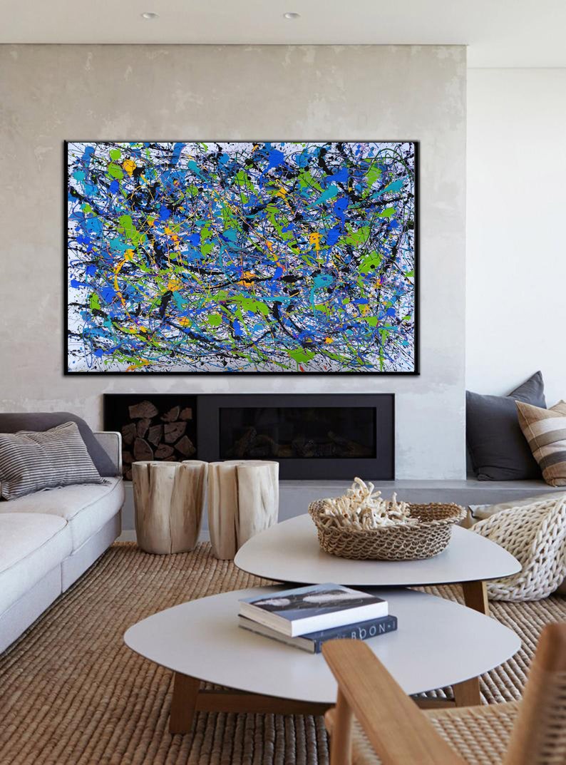 May include: Abstract painting with black, blue, green, and yellow paint splatters on a white background. The painting is framed in black and hanging on a wall above a fireplace.