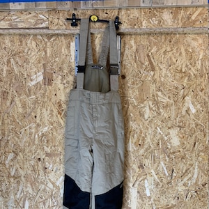May include: Tan snow bib overalls with black reinforced knees hang from a black hanger. The bibs have adjustable straps and pockets. The background is a light brown wood panel.