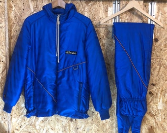 May include: A vintage blue ski suit, comprising a hooded jacket and matching trousers. The jacket has a zip closure, orange accent lines, and the "ellesse" logo. The trousers have an elasticated waistband and cuffs. The suit is displayed on wooden hangers.