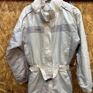 May include: A light beige one-piece ski suit with a high collar and a belt at the waist. The suit has a zipper closure and the brand name "Quechua" is visible on the chest. The suit is hanging on a wooden hanger.