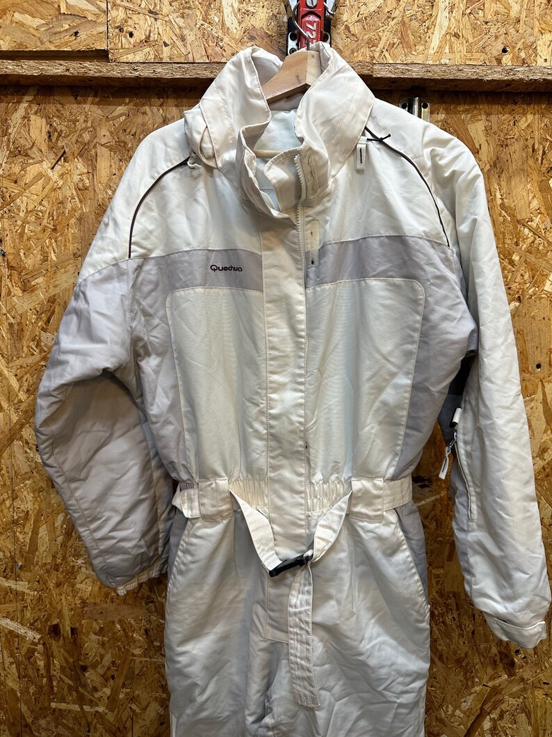 May include: A light beige one-piece ski suit with a high collar and a belt at the waist. The suit has a zipper closure and the brand name "Quechua" is visible on the chest. The suit is hanging on a wooden hanger.