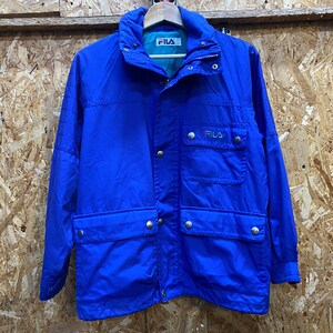 Puede incluir: Chaqueta Fila azul brillante con cuello alto y múltiples bolsillos. La chaqueta tiene cremallera y cierres de botón a presión, y el logotipo de Fila es visible en el pecho. La chaqueta está colgada en una percha de madera.