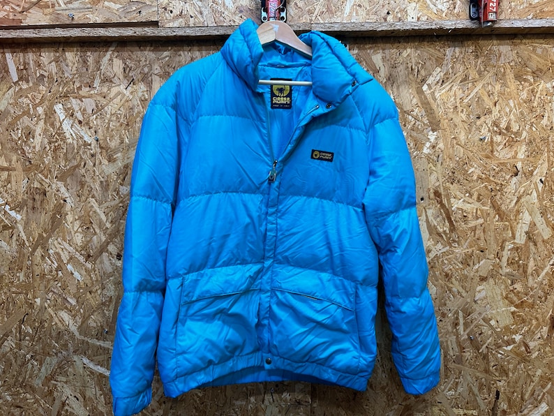 May include: A bright blue puffer jacket with a high collar and a zipper closure. The jacket has a quilted design and a small logo patch on the chest. The jacket is hanging on a wooden hanger against a wood-paneled background.