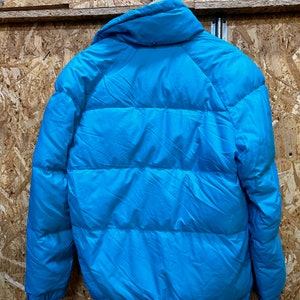 May include: A bright turquoise puffer jacket with a high collar. The jacket is hanging on a wooden surface. The jacket has horizontal quilted lines across the body and sleeves.