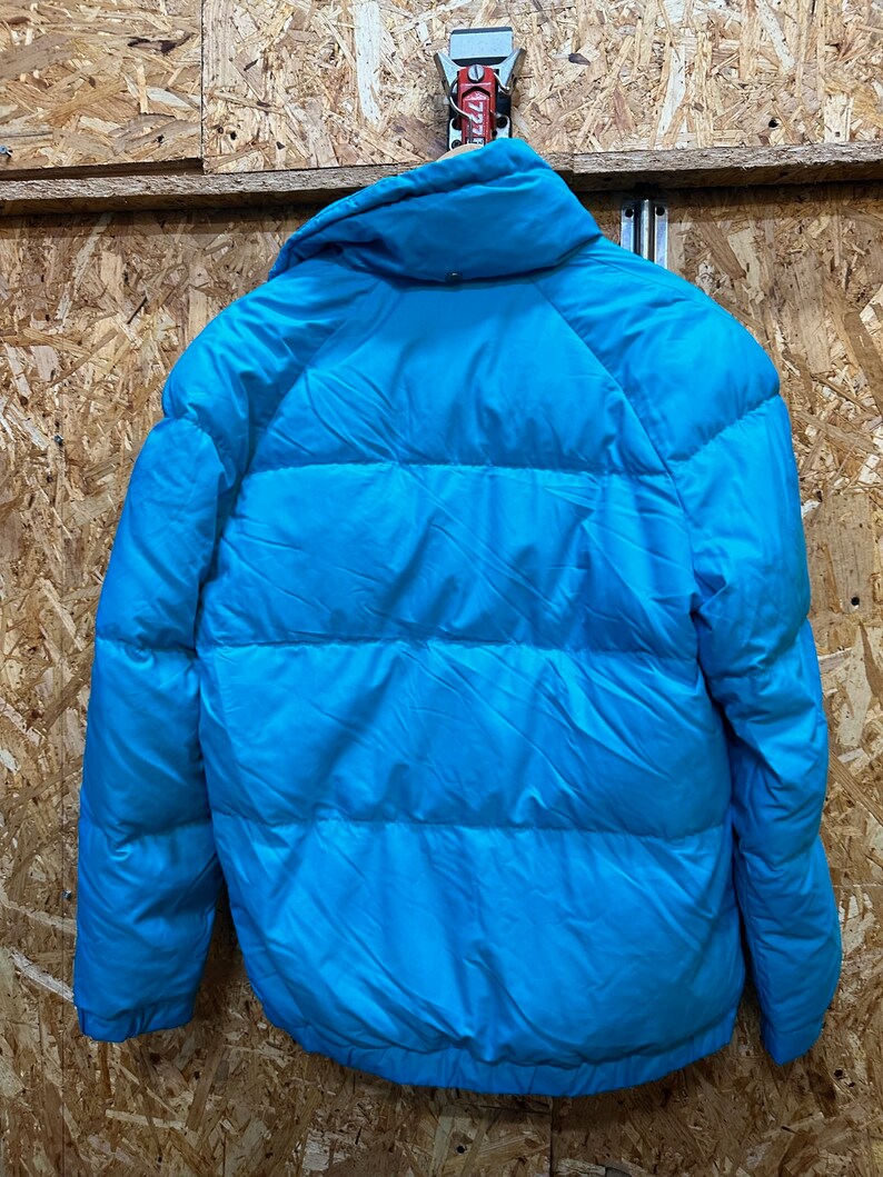 May include: A bright turquoise puffer jacket with a high collar. The jacket is hanging on a wooden surface. The jacket has horizontal quilted lines across the body and sleeves.