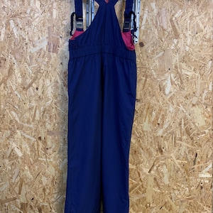 May include: Navy blue snow bib overalls with adjustable straps and a pink interior lining. The bib overalls are hanging on a black hanger against a wood-panelled wall. The overalls have a classic design with a button closure at the ankles.