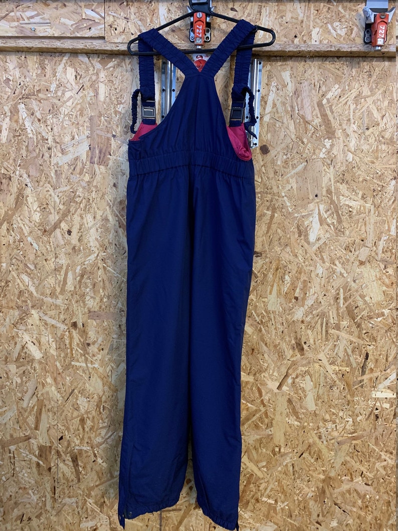 May include: Navy blue snow bib overalls with adjustable straps and a pink interior lining. The bib overalls are hanging on a black hanger against a wood-panelled wall. The overalls have a classic design with a button closure at the ankles.