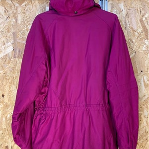 Pode incluir: Casaco com capuz magenta com cintura ajust&aacute;vel por cord&atilde;o e z&iacute;per preto. O casaco est&aacute; pendurado em um cabide preto contra um fundo de madeira marrom claro. Fechamento por bot&atilde;o no capuz.