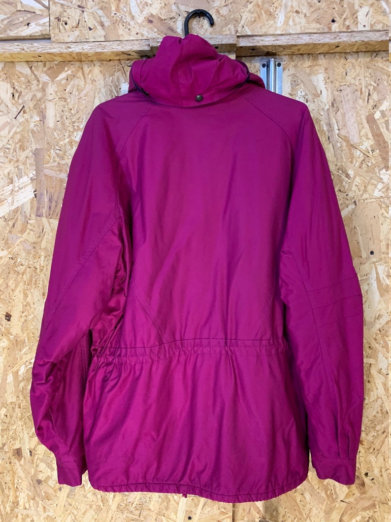 Pode incluir: Casaco com capuz magenta com cintura ajust&aacute;vel por cord&atilde;o e z&iacute;per preto. O casaco est&aacute; pendurado em um cabide preto contra um fundo de madeira marrom claro. Fechamento por bot&atilde;o no capuz.