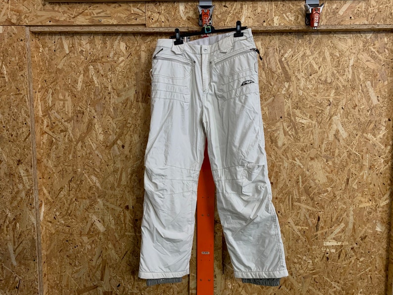 Puede incluir: Pantalones de esqu&iacute; color blanco roto con m&uacute;ltiples bolsillos y rodillas reforzadas colgados de una percha negra. Los pantalones tienen cierre de cremallera y un logotipo en la parte superior de la pierna izquierda. Hechos de material resistente al agua.