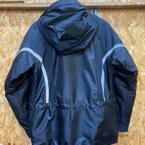 May include: Black hooded jacket with a textured pattern and reflective silver stripes on the sleeves. The jacket has a drawstring waist and is hanging against a wood-paneled wall.