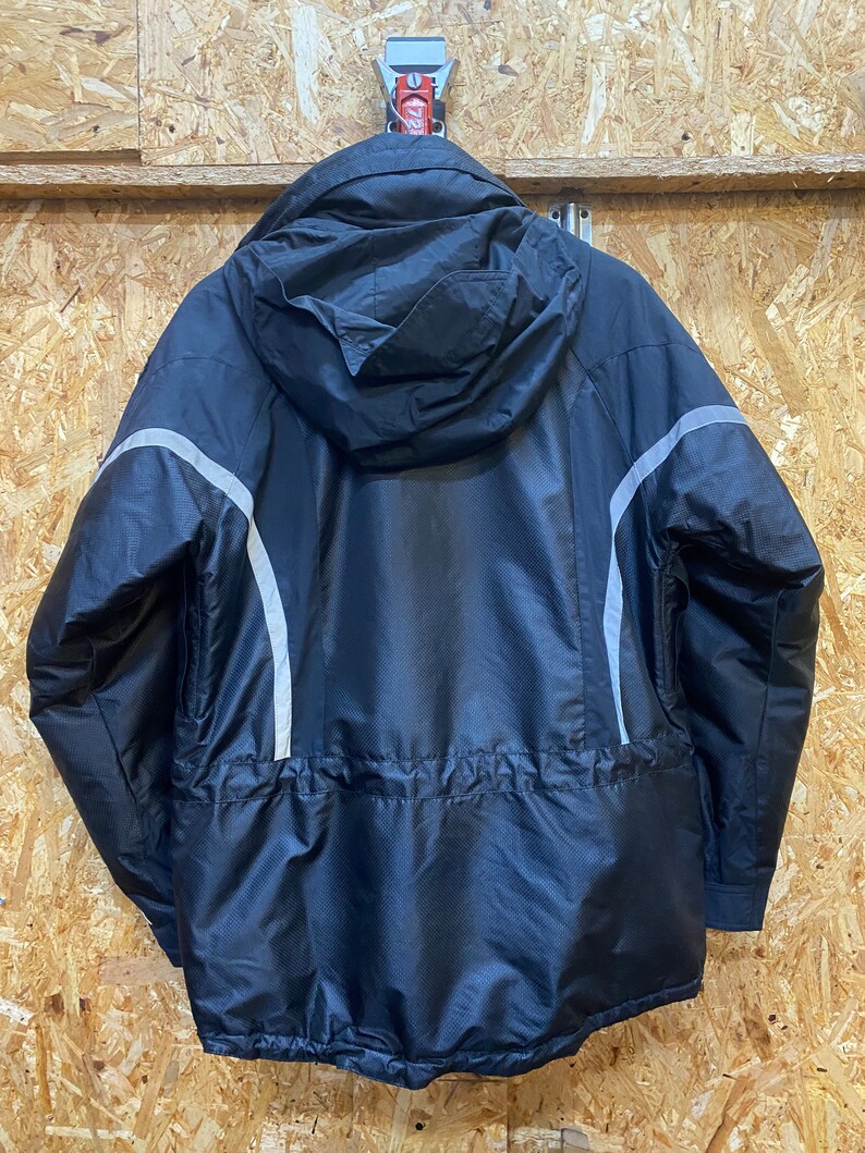 May include: Black hooded jacket with a textured pattern and reflective silver stripes on the sleeves. The jacket has a drawstring waist and is hanging against a wood-paneled wall.