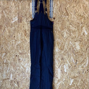 Puede incluir: Mono de esqu&iacute; azul marino con tirantes ajustables y herrajes dorados. El mono tiene un bolsillo delantero con cremallera y est&aacute; colgado de una percha negra contra una pared de paneles de madera. Para deportes de invierno.