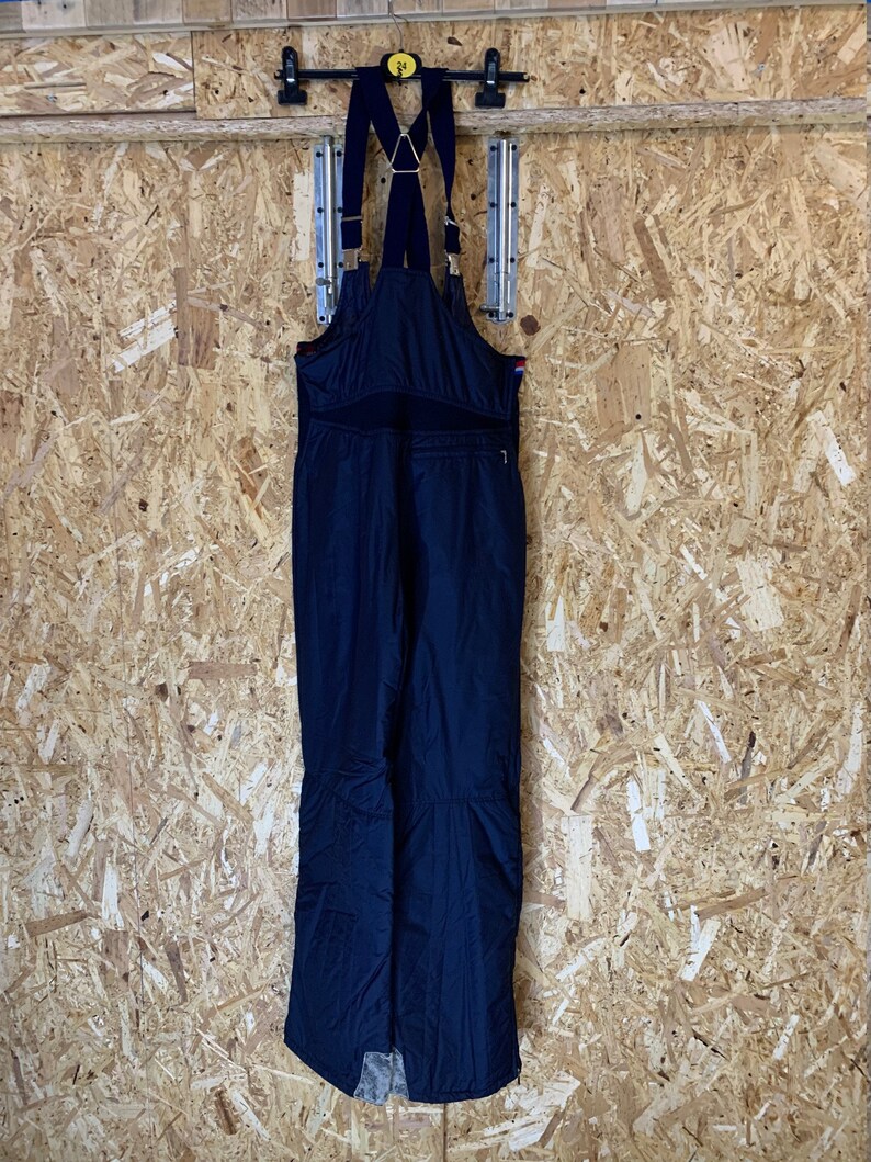 Puede incluir: Mono de esqu&iacute; azul marino con tirantes ajustables y herrajes dorados. El mono tiene un bolsillo delantero con cremallera y est&aacute; colgado de una percha negra contra una pared de paneles de madera. Para deportes de invierno.