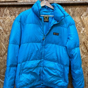 May include: A bright turquoise puffer jacket with a high collar and a full-length zipper. The jacket has the brand name "Esse Piumini" on the chest and a logo on the upper back. The jacket is hanging on a wooden hanger.