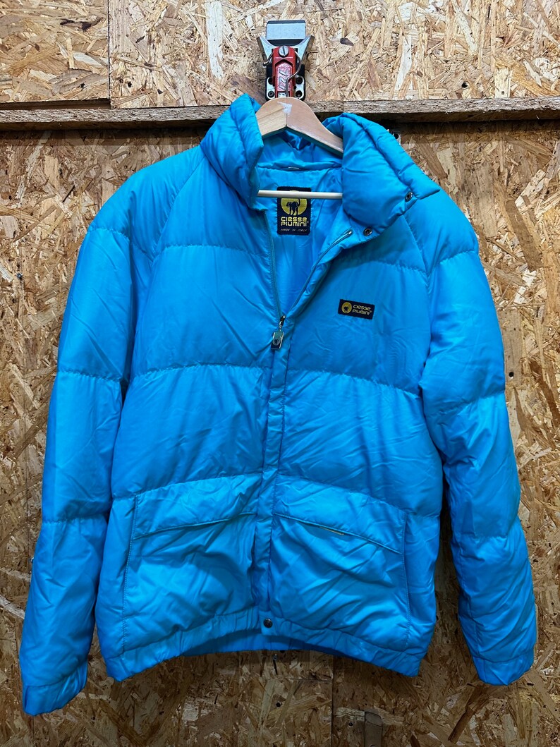 May include: A bright turquoise puffer jacket with a high collar and a full-length zipper. The jacket has the brand name "Esse Piumini" on the chest and a logo on the upper back. The jacket is hanging on a wooden hanger.