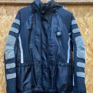 May include: Black Fila jacket with a hood, featuring gray padded accents on the sleeves and shoulders. The jacket has a button-up front, a small logo on the chest, and a reflective stripe down the sleeves.