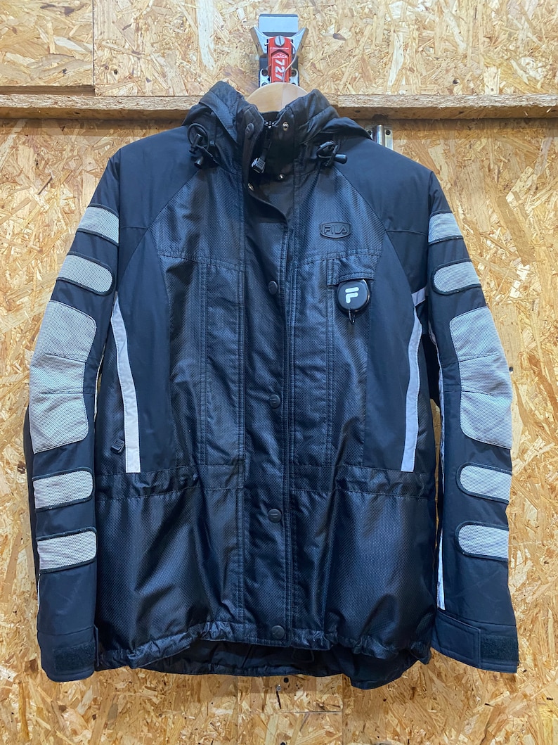 May include: Black Fila jacket with a hood, featuring gray padded accents on the sleeves and shoulders. The jacket has a button-up front, a small logo on the chest, and a reflective stripe down the sleeves.