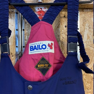 May include: Navy blue and pink ski bib overalls with the brand name "BAILO" and "GORE-TEX" logos. The overalls have adjustable straps and metal button closures. The text "WARRANTED TO BE A BAILO" is visible on a label.