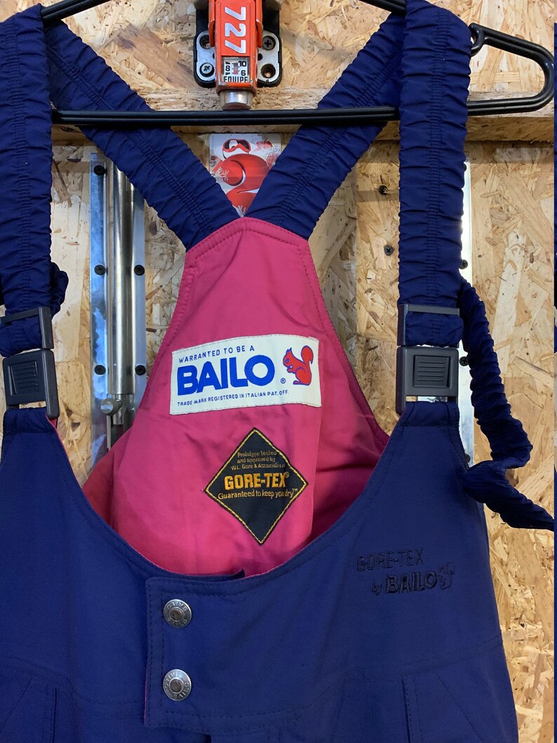 May include: Navy blue and pink ski bib overalls with the brand name "BAILO" and "GORE-TEX" logos. The overalls have adjustable straps and metal button closures. The text "WARRANTED TO BE A BAILO" is visible on a label.