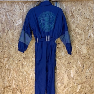 May include: A blue one-piece ski suit with gray accents on the sleeves. The back of the suit features a circular logo with the text "MADE FOR MOTION" and "WFK15". The suit is hanging against a wooden background.