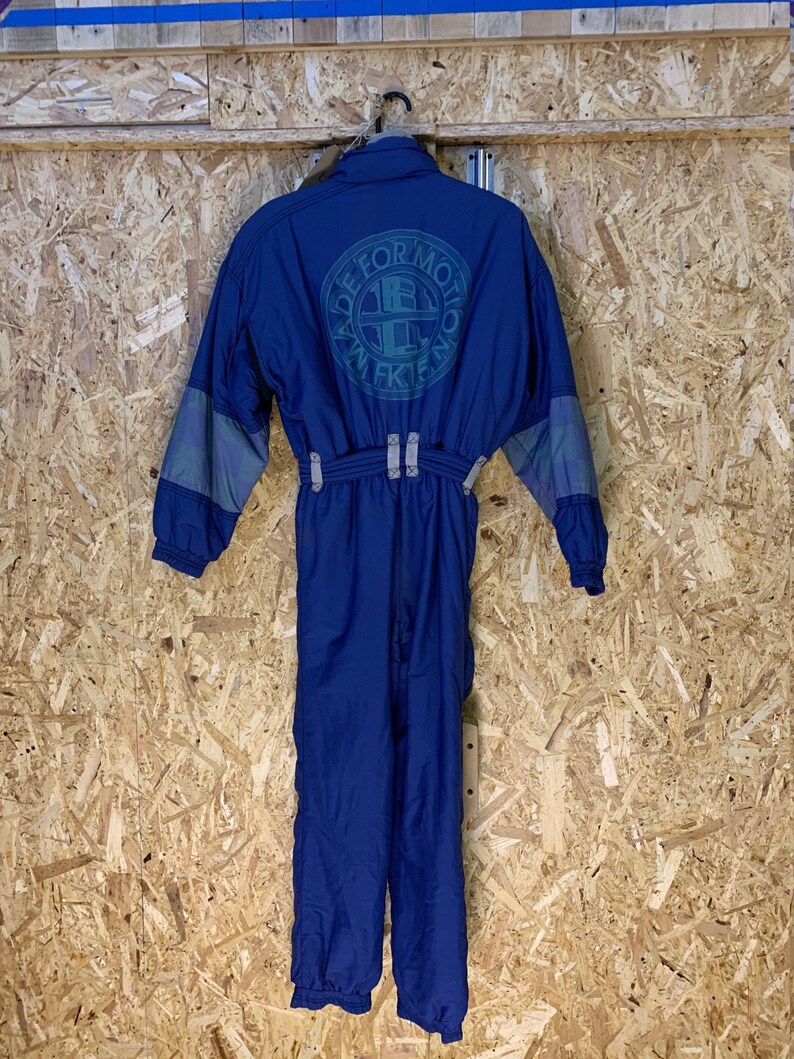 May include: A blue one-piece ski suit with gray accents on the sleeves. The back of the suit features a circular logo with the text "MADE FOR MOTION" and "WFK15". The suit is hanging against a wooden background.