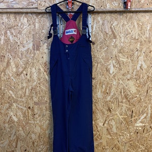 May include: Navy blue ski bib overalls with red detailing and the "BAILO" logo. The overalls feature adjustable straps and a button-up front. The garment is suspended on a black hanger against a wood-panelled wall.