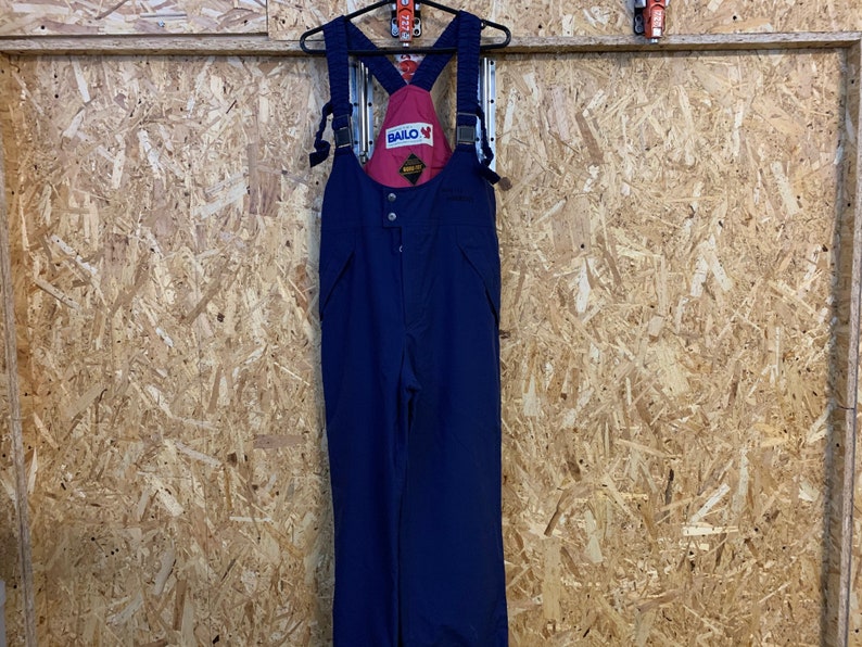 May include: Navy blue ski bib overalls with red detailing and the "BAILO" logo. The overalls feature adjustable straps and a button-up front. The garment is suspended on a black hanger against a wood-panelled wall.