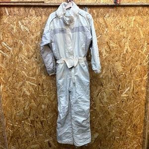 May include: A light-colored one-piece ski suit with a high collar and a belt at the waist. The suit has a zipper closure and the brand name "Quechua" is visible on the chest. The suit is hanging on a wooden hanger.