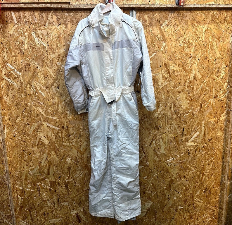 May include: A light-colored one-piece ski suit with a high collar and a belt at the waist. The suit has a zipper closure and the brand name "Quechua" is visible on the chest. The suit is hanging on a wooden hanger.