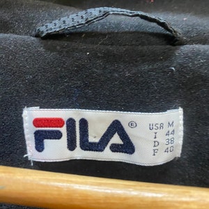 May include: Close-up of a black garment with a label featuring the brand name "FILA" in navy blue and red. The label also includes size information: USA M, I 44, D 38, F 40. The garment has a loop at the top.