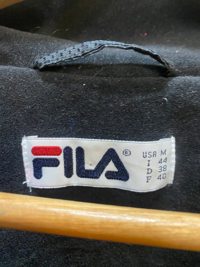 May include: Close-up of a black garment with a label featuring the brand name "FILA" in navy blue and red. The label also includes size information: USA M, I 44, D 38, F 40. The garment has a loop at the top.