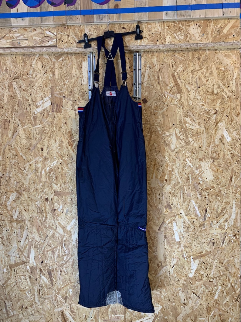 Puede incluir: Mono de esqu&iacute; azul marino con tirantes. El mono tiene un dise&ntilde;o acolchado y detalles de rayas rojas, blancas y azules en las correas de los hombros. El mono est&aacute; colgado de una percha negra sobre un fondo de madera.