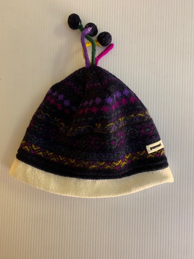 Puede incluir: Un gorro de punto morado oscuro y negro con un borde blanco y un patr&oacute;n colorido. El gorro tiene un dise&ntilde;o &uacute;nico con tres pompones negros en la parte superior, conectados por tallos coloridos. La etiqueta dice "pancari".