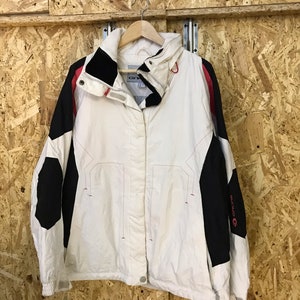 Puede incluir: Chaqueta blanca y negra con detalles rojos, colgada de una percha de madera. La chaqueta tiene un cuello alto y cierre de botones. Las mangas son negras con un logotipo rojo. La chaqueta es de material ligero.